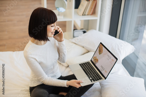 Smiling caucasian woman with brown hair talking on mobile phone and browsing online shop with fast food on laptop. Ordering of grocery delivery during sale season. Modern lifestyles with gadgets.