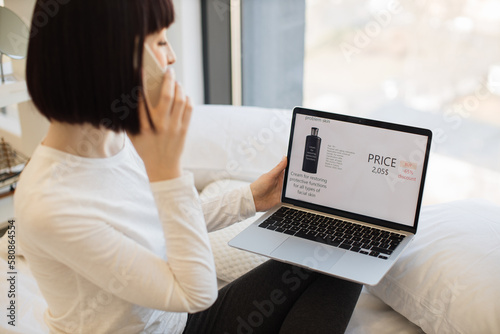 Happy caucasian woman resting on bed with wireless laptop and using smartphone for ordering cosmetic during sale season. Discount of product with 65 percent in online shop.