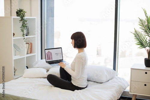 Back view of darked haired woman sitting on white bed and browsing website with trendy clothes on sale. Caucasian female using wireless laptop for choosing new wear with discount of 25 percent online.