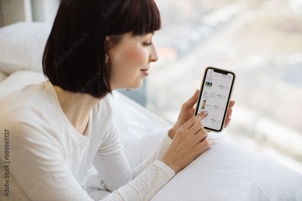 Happy charming lady with brown hair choosing professional cosmetics on sale while browsing website. Caucasian woman in casual attire using modern smartphone for online shopping at cozy home. Happy charming lady with brown hair choosing professional cosmetics on sale while browsing website. Caucasian woman in casual attire using modern smartphone for online shopping at cozy home.