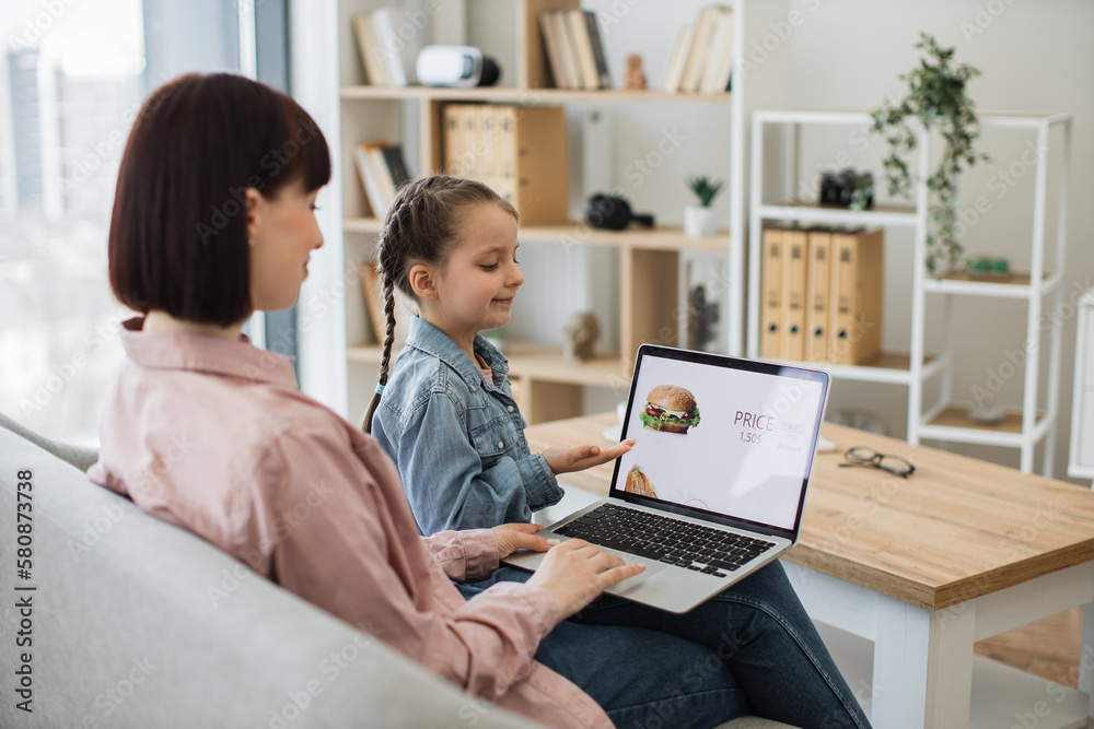 Attractive dark haired woman sitting on couch with her lovely daughter and choosing fast food with discount on website. Caucasian family of two using modern laptop for ordering takeaway lunch. Attractive dark haired woman sitting on couch with her lovely daughter and choosing fast food with discount on website. Caucasian family of two using modern laptop for ordering takeaway lunch.