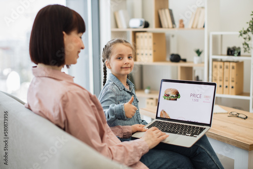 Side view of charming brunette sitting on couch with daughter and using wireless laptop. Girl smiling and looking at camera, showing thumb up while focused mother scrolling online shop with fast food.