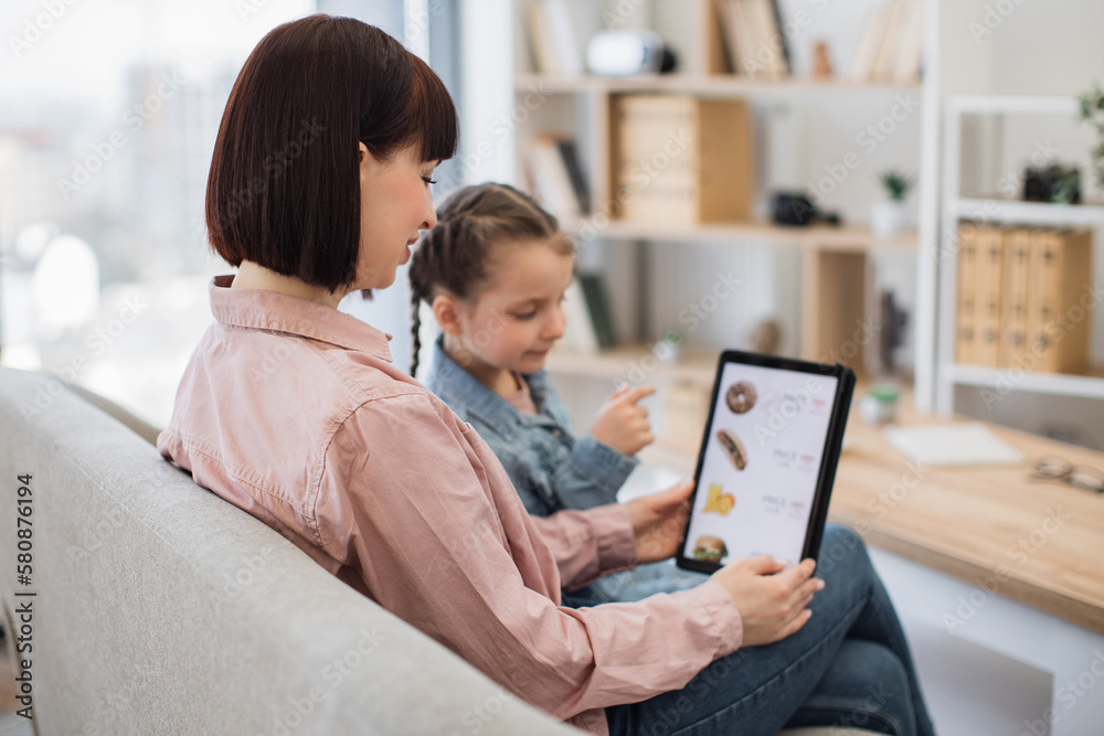 Close up of smiling shoppers choosing fast food at discount on delivery webpage while relaxing at home. Serene woman and girl spending quality time together while buying precooked meals via tablet. Close up of smiling shoppers choosing fast food at discount on delivery webpage while relaxing at home. Serene woman and girl spending quality time together while buying precooked meals via tablet.