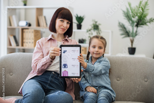 Smiling beautiful lady with pretty child holding tablet in hands with discount offer for clothes on screen. Happy caucasian females enjoying e-commerce while browsing online fashion store indoors. photo