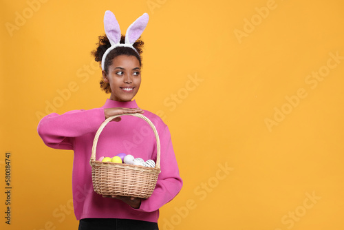 Happy African American woman in bunny ears headband holding wicker basket with Easter eggs on orange background, space for text