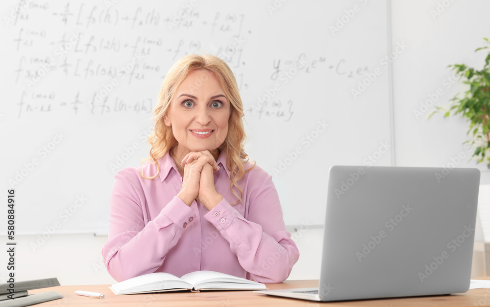 Happy teacher sitting near laptop at desk in classroom Happy teacher sitting near laptop at desk in classroom