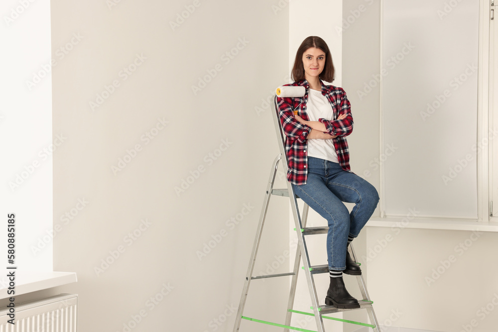 Young beautiful woman with roller on metal stepladder indoors. Room renovation