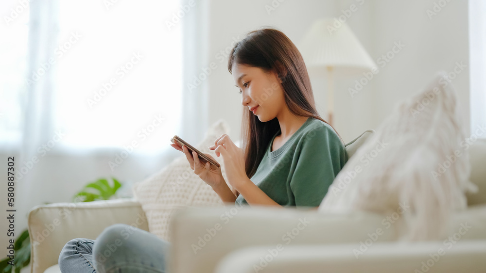 Happy young asian woman relax on comfortable couch at home texting messaging on smartphone, smiling girl use cellphone chatting, browse wireless internet on gadget, shopping online from home - obrazy, fototapety, plakaty 