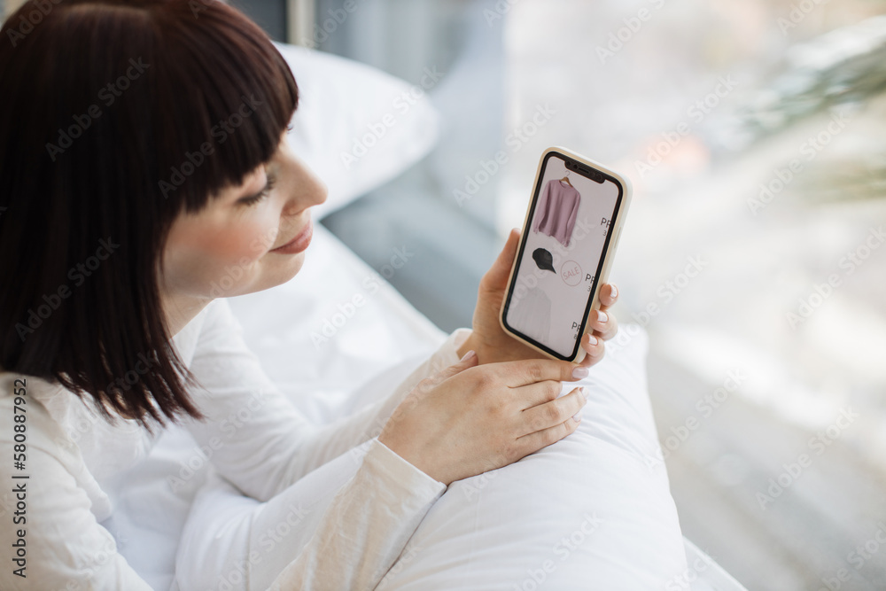 Pleasant caucasian woman in casual outfit using personal smartphone for ordering new trendy clothes online while resting at home. Shopping during sale season with help of modern gadgets.