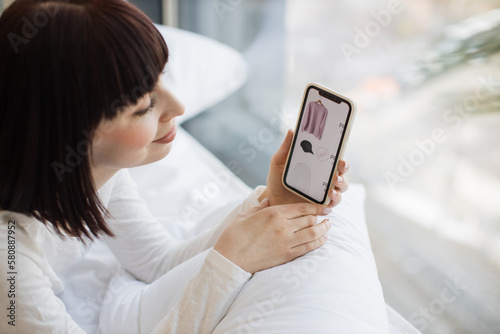 Pleasant caucasian woman in casual outfit using personal smartphone for ordering new trendy clothes online while resting at home. Shopping during sale season with help of modern gadgets.