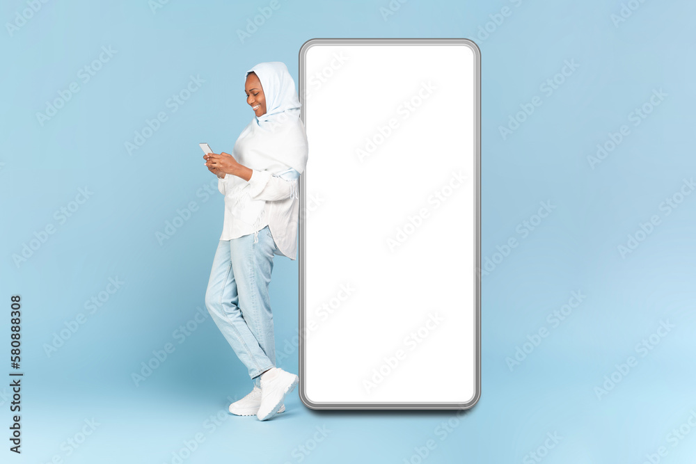 Online offer concept. Happy african american woman in hijab using cellphone, leaning on big phone with empty screen Online offer concept. Happy african american woman in hijab using cellphone, leaning on big phone with empty screen