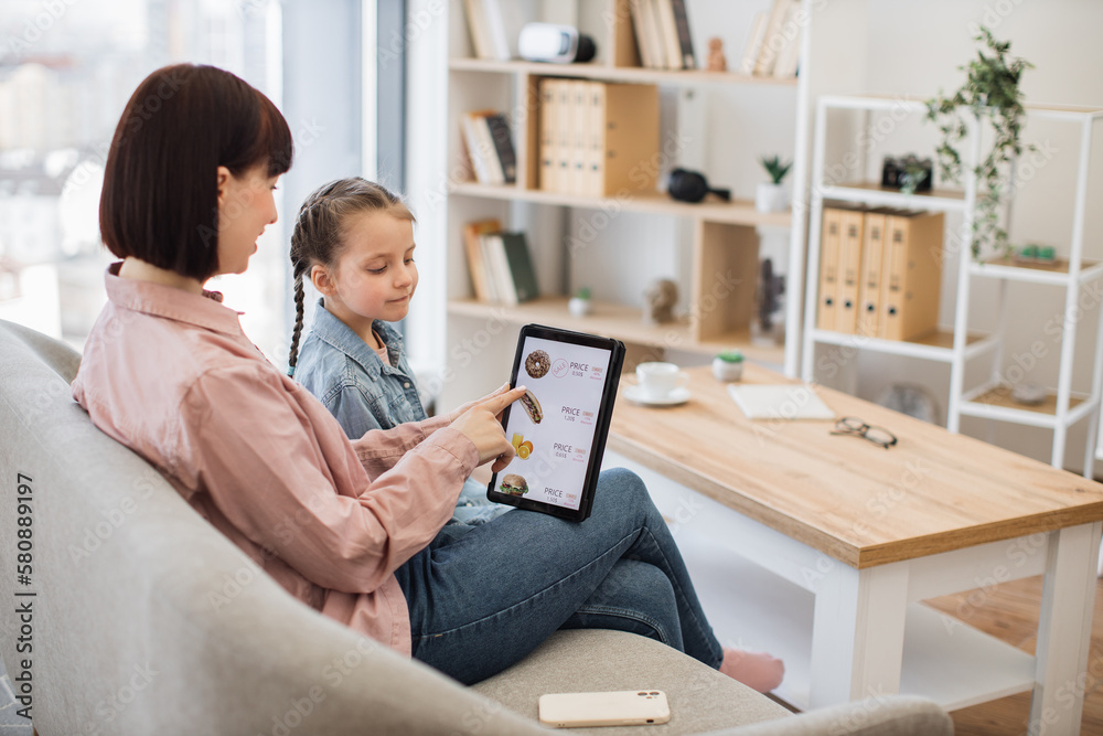 Relaxed mother and daughter searching e-commerce offers on restaurant food with tablet application while having fun indoors. Efficient online clients seizing moment of reduced prices on products. Relaxed mother and daughter searching e-commerce offers on restaurant food with tablet application while having fun indoors. Efficient online clients seizing moment of reduced prices on products.