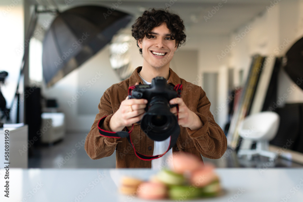 Young male food photographer using digital camera and taking photo of dessert, looking and smiling at camera