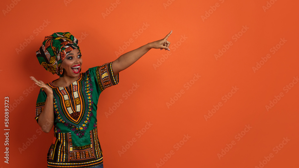 Emotional black lady in african costume pointing at copy space
