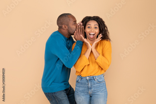 Middle aged man sharing secrets with his surprised wife, whispering to her ear, standing over beige background