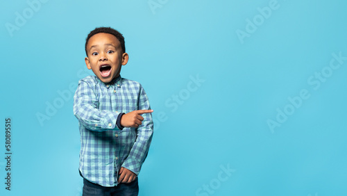 Special offer. Adorable african american boy pointing finger aside at copy space and looking at camera with open mouth