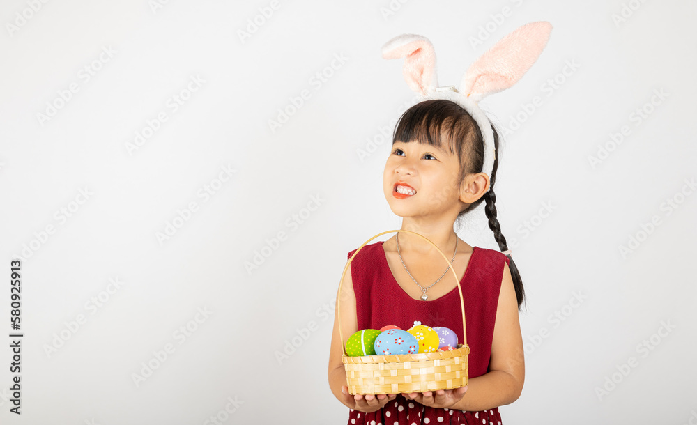 Happy Easter Day. Smile Asian little girl wearing easter bunny ears holding basket of full colorful eggs smiles broadly isolated on white background with copy space, Happy child in holiday Happy Easter Day. Smile Asian little girl wearing easter bunny ears holding basket of full colorful eggs smiles broadly isolated on white background with copy space, Happy child in holiday