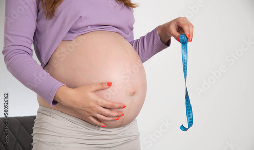 A pregnant girl with a big belly holds a measuring tape on a white background. The concept of measurement and size of the abdomen during pregnancy. The volume of the abdomen by week. photo