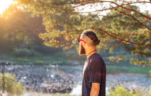 Handsome perfect man with a beard and black glasses outdoors in summer against the backdrop of a forest and a river bank. The concept of outdoor recreation, survival in nature. Bushcrafting.  © HENADZY