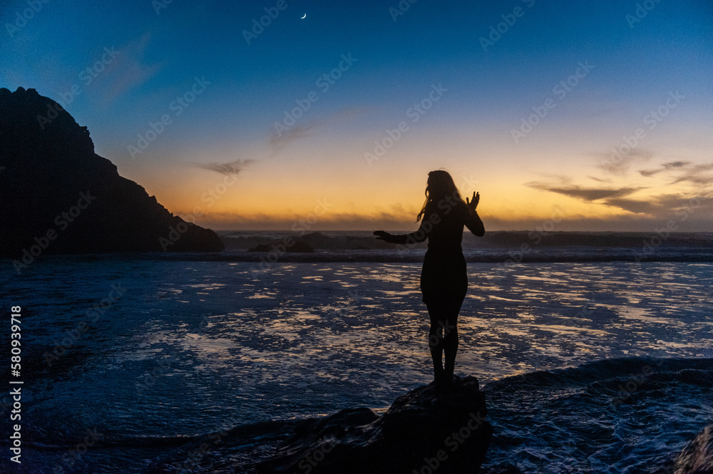 Silhoutte of a young woman contrasted against the orange sunset glow, with a crescent moon in the background.