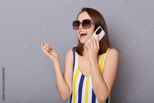Portrait of excited happy woman with brown hair wearing striped colorful swimming suit and sunglasses, talking on mobile phone with toothy smile isolated on gray background