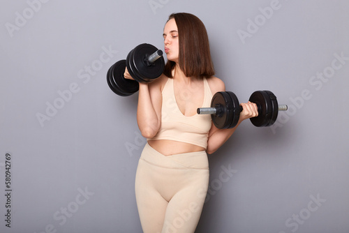 Indoor shot attractive athletic woman wearing sportswear working out with dumbbells isolated on gray background, kissing barbel, enjoying her workout, training her biceps and triceps