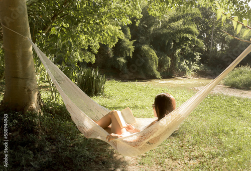 Woman reading book while relaxing on hammock in park during sunny day