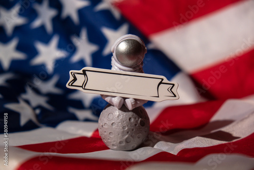 Plastic toy figure astronaut with paper note for your text on American flag background Copy space. 50th Anniversary of USA Landing on The Moon Concept of out of earth travel, private spaceman 