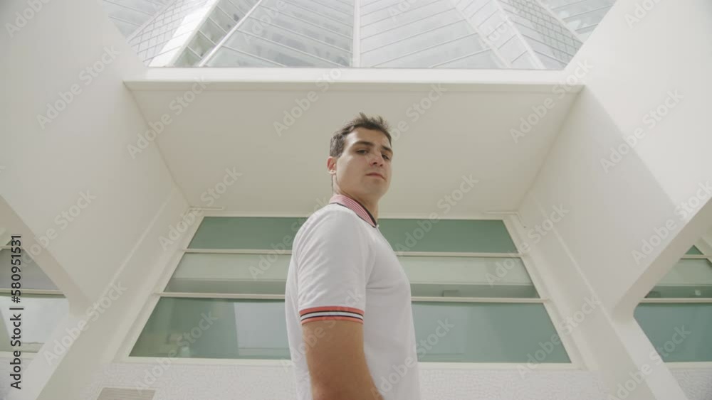 Low angle view of a teenager in white t-shirt looking at camera. Close-up shot of a confident and serious guy in his 20s within modern interior. High quality 4k footage Low angle view of a teenager in white t-shirt looking at camera. Close-up shot of a confident and serious guy in his 20s within modern interior. High quality 4k footage