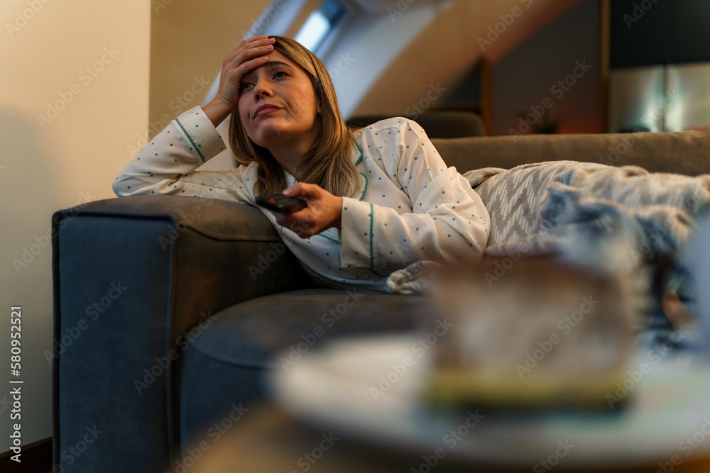 Photo of girl laying on a sofa and using remote control while watching TV, she looks bored