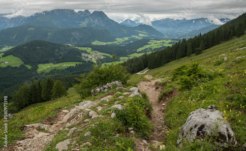 Hiking path between green grassy alpine slopes viewing towards pine trees and a beautiful rocky peak in Austria during a summer day.