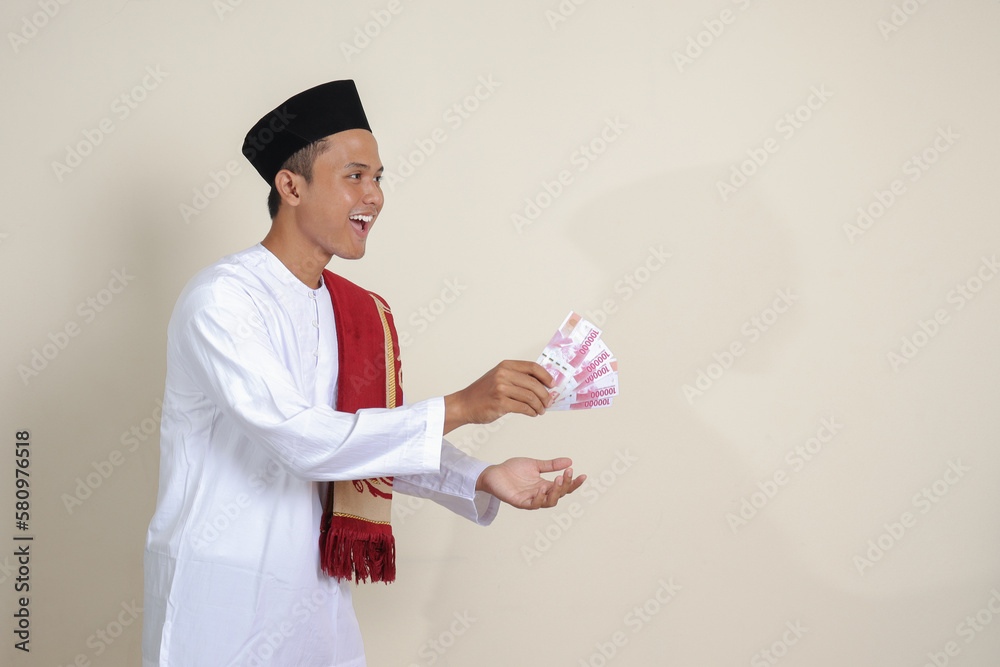 Portrait of attractive Asian muslim man in white shirt with skullcap giving one hundred thousand rupiah for charity. Isolated image on gray background Portrait of attractive Asian muslim man in white shirt with skullcap giving one hundred thousand rupiah for charity. Isolated image on gray background
