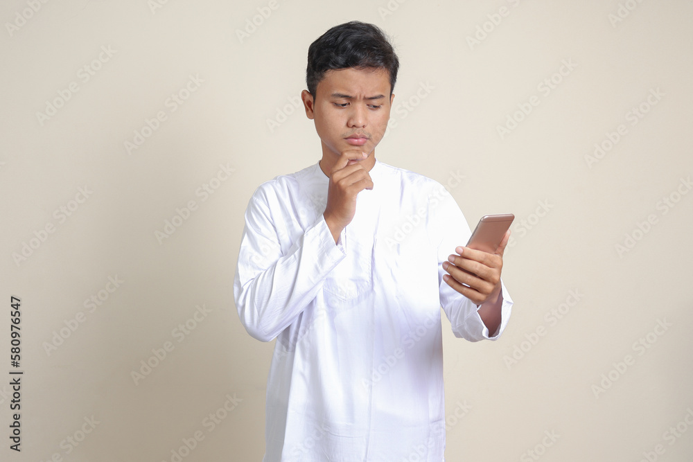 Portrait of attractive Asian muslim man in white shirt thinking about question with hand on chin while holding a mobile phone. Isolated image on gray background Portrait of attractive Asian muslim man in white shirt thinking about question with hand on chin while holding a mobile phone. Isolated image on gray background