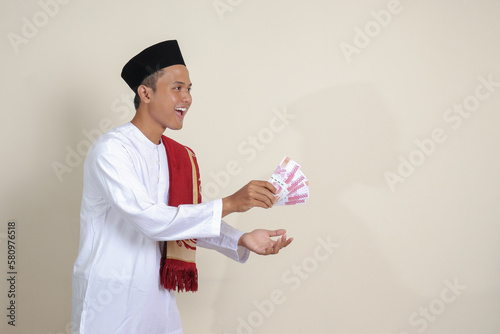 Portrait of attractive Asian muslim man in white shirt with skullcap giving one hundred thousand rupiah for charity. Isolated image on gray background