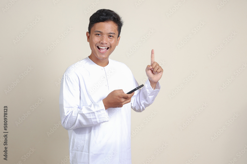 Portrait of attractive Asian muslim man in white shirt holding mobile phone with smiling expression on face while pointing finger to the side. Advertising concept. Isolated image on gray background Portrait of attractive Asian muslim man in white shirt holding mobile phone with smiling expression on face while pointing finger to the side. Advertising concept. Isolated image on gray background