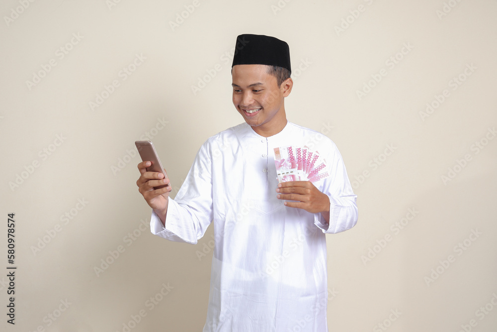 Portrait of attractive Asian muslim man in white shirt showing one hundred thousand rupiah while using mobile phone. Financial and savings concept. Isolated image on gray background Portrait of attractive Asian muslim man in white shirt showing one hundred thousand rupiah while using mobile phone. Financial and savings concept. Isolated image on gray background