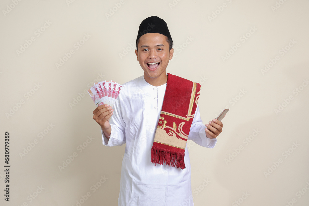 Portrait of attractive Asian muslim man in white shirt showing one hundred thousand rupiah while using mobile phone. Financial and savings concept. Isolated image on gray background