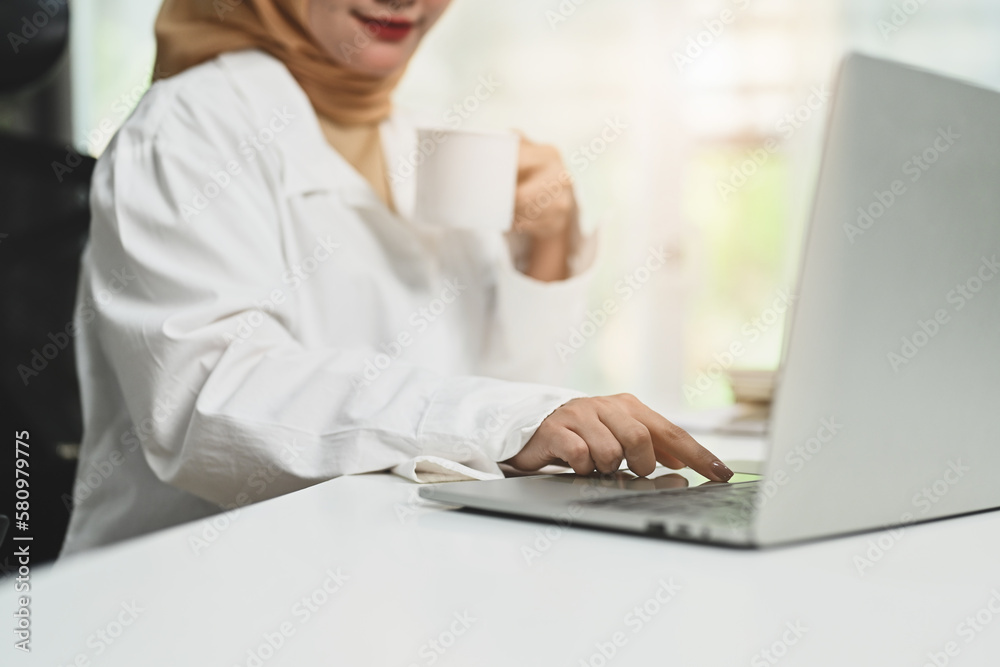 Cropped shot of smiling young muslim woman drinking coffee and checking email on laptop at her office desk Cropped shot of smiling young muslim woman drinking coffee and checking email on laptop at her office desk