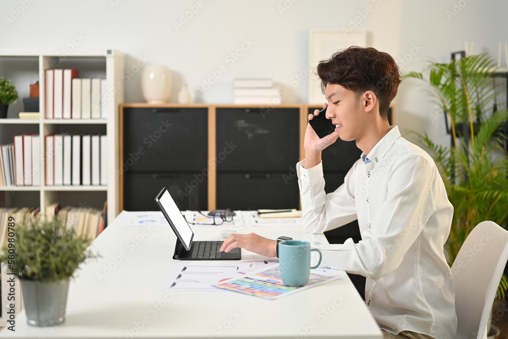Side view of young creative man in white shirt using digital tablet and talking on mobile phone Side view of young creative man in white shirt using digital tablet and talking on mobile phone
