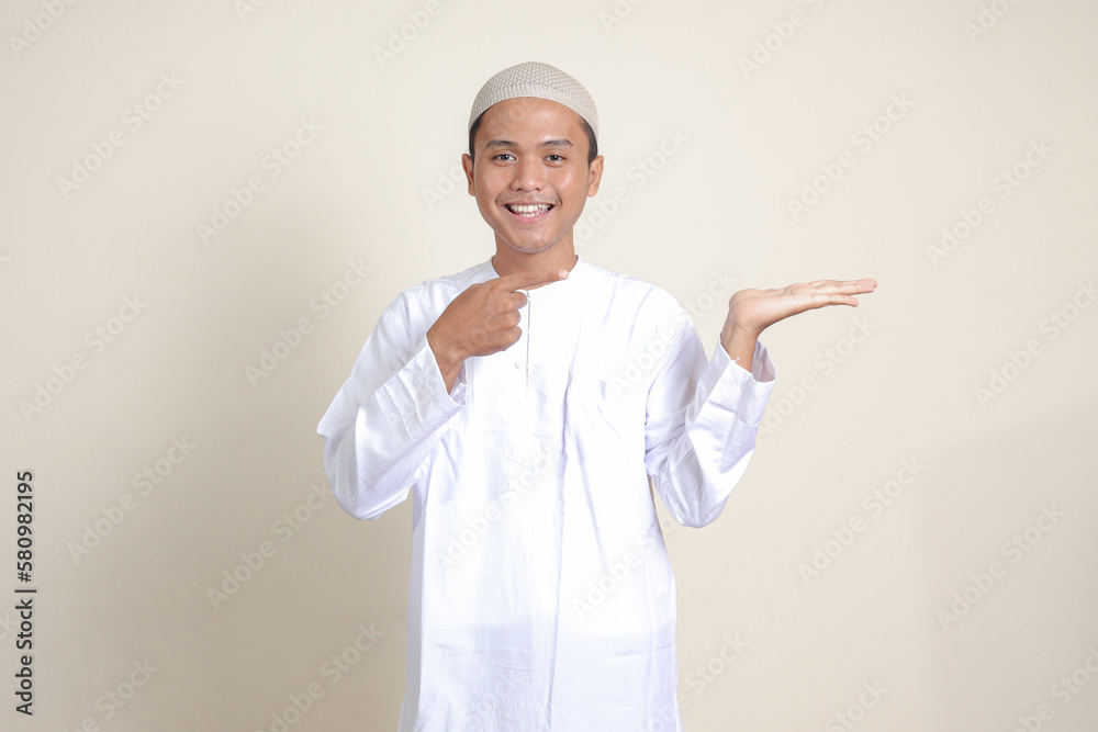 Portrait of attractive Asian muslim man in white shirt with skullcap showing product and pointing with his hand and finger to the side. Advertising concept. Isolated image on gray background Portrait of attractive Asian muslim man in white shirt with skullcap showing product and pointing with his hand and finger to the side. Advertising concept. Isolated image on gray background