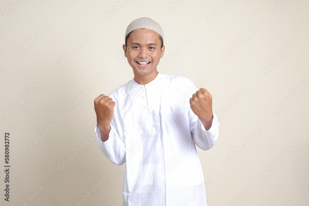 Portrait of attractive Asian muslim man in white shirt raising his fist, celebrating success. Isolated image on gray background Portrait of attractive Asian muslim man in white shirt raising his fist, celebrating success. Isolated image on gray background