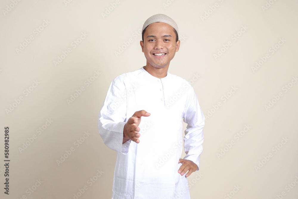 Portrait of attractive Asian muslim man in white shirt reaching out his hand for a handshake, welcoming someone. Isolated image on gray background