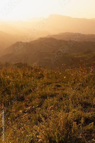 Idyllic landscape - silhouette of high mountain orange ridges in golden yellow pastel sunset light away, mist, grass, flowers on meadow in summer evening, panorama view. Majestic travel in Dagestan.