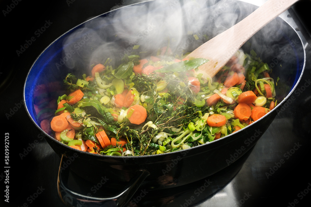 Steaming vegetables and herbs in a cooking pan on the black stove top, ingredients for a soup like carrots, celery, leek, thyme and bay leaves, copy space, selected focus Steaming vegetables and herbs in a cooking pan on the black stove top, ingredients for a soup like carrots, celery, leek, thyme and bay leaves, copy space, selected focus
