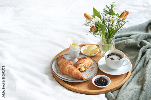 Sunday breakfast in bed with coffee, croissant, jam and boiled egg served on a wooden tray with a bouquet of flowers on mother's day, birthday or valentine's day, copy space