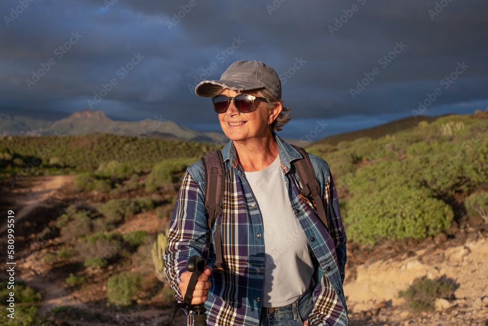 Beautiful senior woman in hat and sport clothing in trekking day walking at sunset enjoying freedom and vacation. Happy mature female with stick looking the mountain tropical landscape