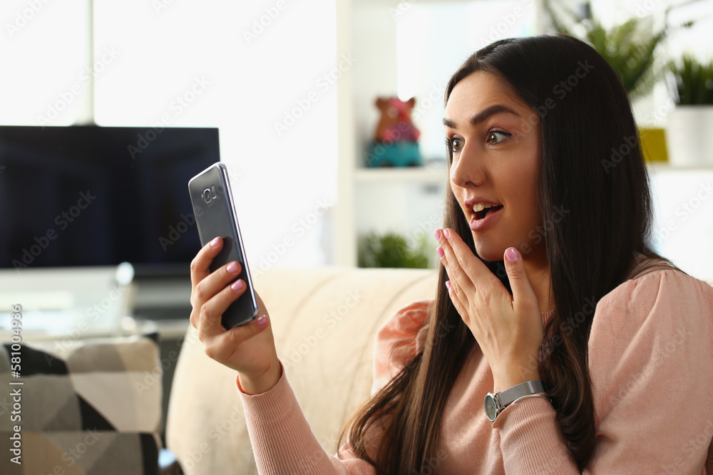 Woman surprised in shock looks at smartphone screen while sitting on couch at home Woman surprised in shock looks at smartphone screen while sitting on couch at home