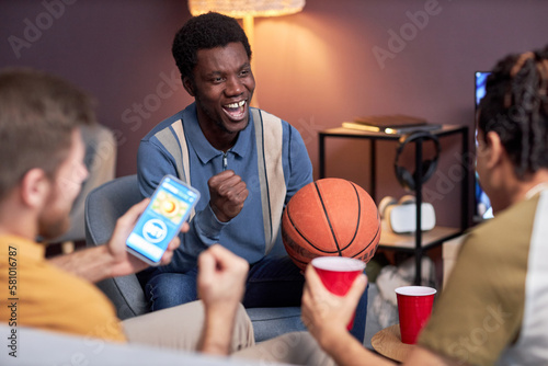 Portrait of emotional black man cheering for basketball match at home with friends and holding ball © Seventyfour