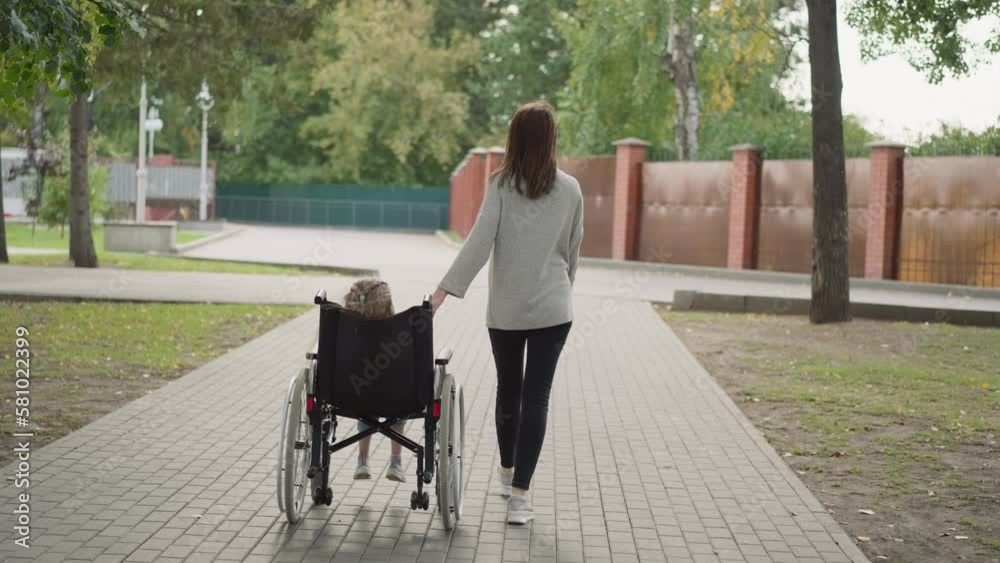 Mother helps little daughter to move in wheelchair on path in city park. Woman and girl with spinal cord injury enjoy walk on spring day backside view Mother helps little daughter to move in wheelchair on path in city park. Woman and girl with spinal cord injury enjoy walk on spring day backside view