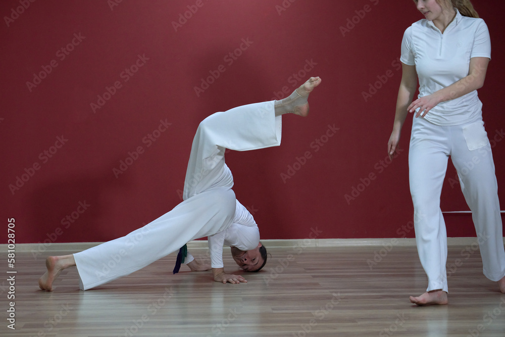 A mature man, a sports master, and a young woman, an aspiring athlete, practice the Brazilian martial art of capoeira in a small gym.